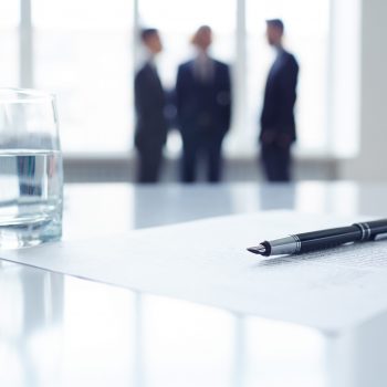 Image of business document, pen and glass of water at workplace with group of colleagues on background